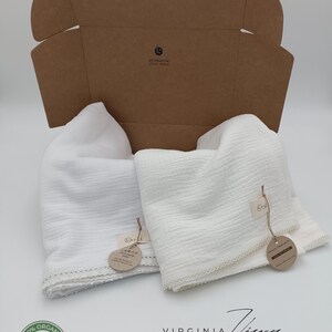 May include: Two white muslin swaddles with lace trim, packaged in a brown cardboard box. The box has a label that says "For little people". The swaddles have tags that say "100% Organic Cotton" and "Virginia Ziggy Art Studio".