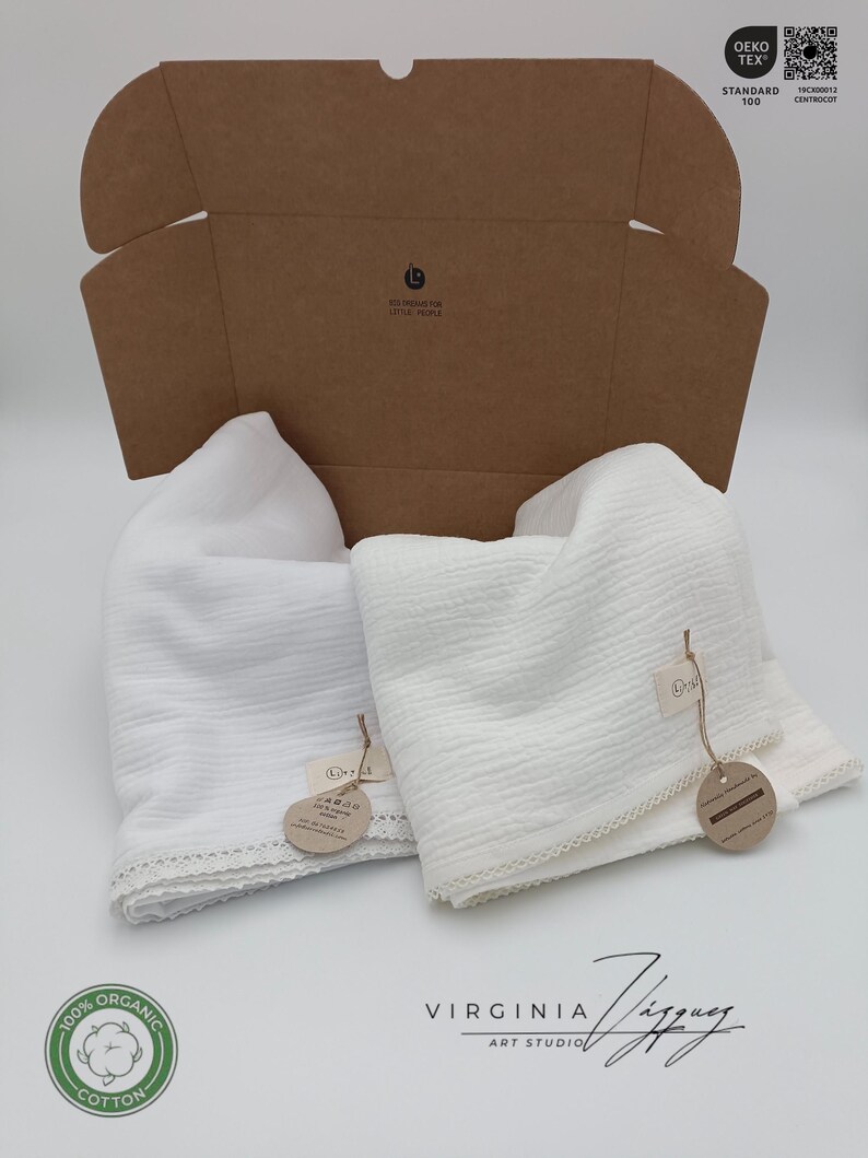 May include: Two white muslin swaddles with lace trim, packaged in a brown cardboard box. The box has a label that says "For little people". The swaddles have tags that say "100% Organic Cotton" and "Virginia Ziggy Art Studio".
