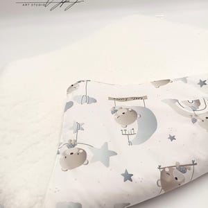 May include: A white baby blanket with a soft, plush texture. The blanket features a whimsical design of brown bears hanging from stars and a crescent moon. The fabric is printed with a subtle gray polka dot pattern. The blanket is certified Oeko-Tex Standard 100.