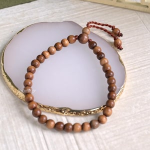 May include: A wooden bead bracelet with a brown cord closure. The bracelet features multiple small, round, light brown beads. The cord is a darker brown color. The bracelet is displayed on a white and gold decorative plate.