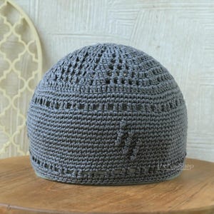 May include: A gray crocheted skullcap with a rounded crown and openwork design. The cap is made of a soft, textured yarn and has a slightly domed shape. The cap is sitting on a wooden surface.