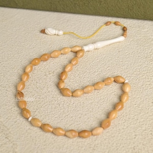 May include: A light brown prayer bead necklace with oval-shaped beads. The beads are strung together with a yellow cord and have a white handle. The necklace is likely used for prayer or meditation.