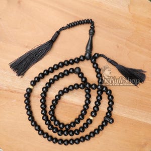 May include: Black prayer beads with a tassel and a wooden handle. The beads are round and smooth, with a few lighter-colored beads interspersed. The image includes the text "The Kufi Shop: Handcrafted Kufis and Misbahas."