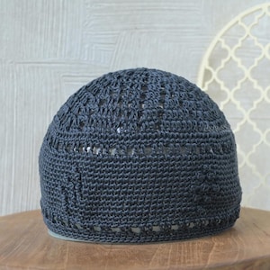 Puede incluir: Un gorro tejido de color gris oscuro con un diseño de tejido abierto. El gorro tiene una corona redondeada y un patrón decorativo cerca de la base. El sombrero está hecho de un material suave y flexible y es adecuado para uso religioso o cultural.