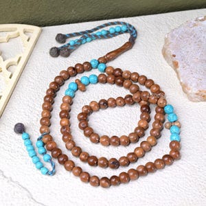 May include: A prayer bead necklace with brown wooden beads and turquoise accents. The beads are strung on a brown and blue cord, with a tassel at the end. The necklace is displayed on a white surface.