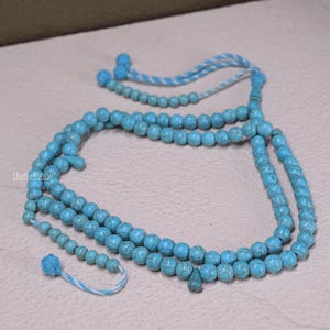 May include: A turquoise prayer bead necklace with multiple strands of round beads. The beads are a light blue color with a marbled texture. The necklace has a tassel and a decorative bead at the end of the strands. The text "TheKuffShop" is visible.