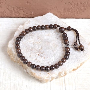 May include: A dark brown beaded bracelet with a knotted closure. The bracelet is made of small, round wooden beads. The beads are strung together on a dark brown cord. The bracelet is displayed on a white stone surface.