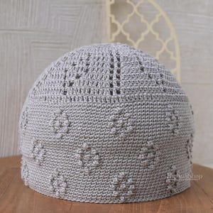 May include: A light gray crocheted skullcap with a rounded crown. The cap features a textured pattern of raised floral designs and openwork details. The handmade hat is displayed on a wooden surface.