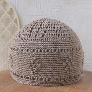 Puede incluir: Un gorro de calavera de ganchillo color topo con un diseño calado. El gorro presenta un patrón texturizado y una corona redondeada. La tela de ganchillo es de un color neutro, adecuado para varios atuendos. El gorro es probablemente hecho a mano.