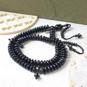 Op de afbeelding: Een zwarte gebedsketting met een kwast en decoratieve kralen. De kralen zijn rond en glad, en de ketting is gemaakt van een donker, mogelijk houten materiaal. De ketting wordt gepresenteerd op een wit oppervlak.