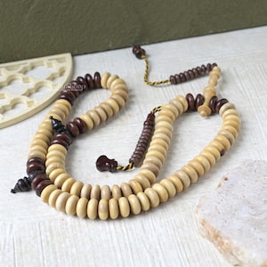 May include: A wooden prayer bead necklace with alternating light and dark brown beads. The beads are strung together with a yellow and black cord. The necklace is displayed on a light-colored wooden surface.