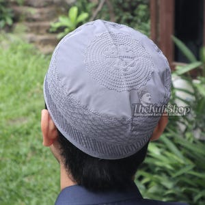 May include: A gray kufi hat with intricate embroidered detailing. The hat features a circular design on top and a textured band around the base. The Kufi Shop logo is visible on the side.