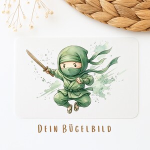 Könnte beinhalten: Rechteckige Karte mit einer Aquarellillustration eines Cartoon-Ninjas in grünem Outfit, der ein Holzschwert hält. Der Text "DEIN BÜGELBILD" ist unten in Braun gedruckt. Der Hintergrund hat grüne Farbspritzer.