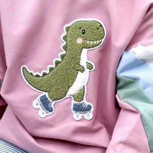 Könnte beinhalten: Ein altrosa Langarm-Sweatshirt mit einer Applikation eines texturierten grünen Dinosauriers, der blaue Rollschuhe trägt. Die Ärmel sind mit farbigen Streifen in Weiß, Hellblau und Salbeigrün abgesetzt. Das Sweatshirt hat einen Rundhalsausschnitt.