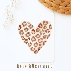 Könnte beinhalten: Eine weiße Karte mit einem herzförmigen Design mit Leopardenmuster in Braun- und Rosatönen. Der Text "DEIN BÜGELBILD" ist unten in Braun gedruckt. Ein Zweig getrockneter Blumen und eine gewebte Matte befinden sich im Hintergrund.