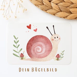Könnte beinhalten: Rechteckige Karte mit einer Aquarellillustration einer lächelnden Schnecke mit rosa Gehäuse, Blättern, Pilzen und Herzen. Der Text "DEIN BÜGELBILD" ist unten in Braun gedruckt. Die Karte liegt auf einer weißen Oberfläche.
