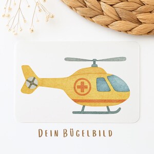 Könnte beinhalten: Illustration eines gelb-orangen Hubschraubers im Aquarellstil mit einem roten Kreuzsymbol auf einer weißen Karte. Die Karte trägt unten den Text "DEIN BÜGELBILD" in braunen Buchstaben. Der Hintergrund umfasst einen geflochtenen Korb und Trockenblumen.