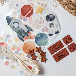 Könnte beinhalten: Eine Sammlung von Bastelmaterialien zum Thema Weltraum. Enthält eine Rakete, Planeten, Sterne, einen Astronauten, hölzerne sternförmige Knöpfe und Etiketten mit deutschem Text. Ein Band mit dem Text "YAY!" ist ebenfalls vorhanden.