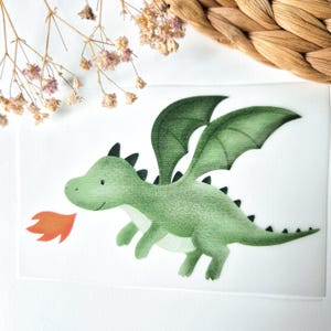 Könnte beinhalten: Aquarell-Illustration eines freundlichen grünen Drachen mit großen Flügeln und schwarzen Stacheln. Der Drache speit eine kleine orangefarbene Flamme. Das Bild befindet sich auf weißem Hintergrund, mit getrockneten Blumen und gewebtem Material in den Ecken.