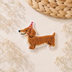 Könnte beinhalten: Ein gestickter Aufnäher in Form eines braun-cremefarbenen Dackels mit strukturierter, flauschiger Oberfläche. Der Hund trägt einen kleinen, spitzen Partyhut mit rosa, gelben und orangefarbenen Streifen. Der Aufnäher hat einen weißen Rand.