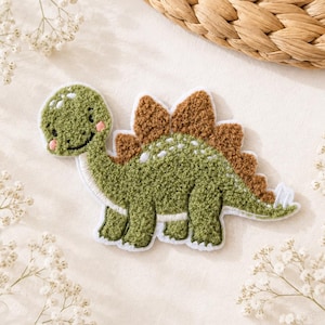 Könnte beinhalten: Ein grüner Dinosaurier-Aufnäher mit einer strukturierten, flauschigen Oberfläche. Der Dinosaurier hat braune Platten auf dem Rücken, ein lächelndes Gesicht mit rosa Wangen und einen weißen Rand. Das Design ist in einem weichen Stickstil gehalten.