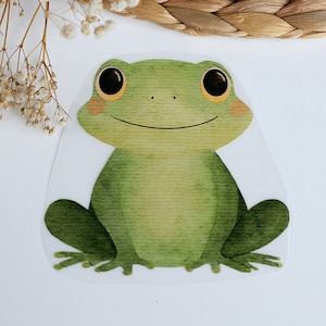 Könnte beinhalten: Ein grüner Frosch-Aufkleber mit Aquarell-Effekt. Der Frosch hat große schwarze Augen mit gelben Highlights, rosige Wangen und einen lächelnden Mund. Der Aufkleber befindet sich auf weißem Hintergrund.