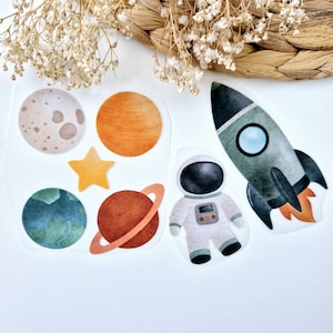 Könnte beinhalten: Ein Set bunter Weltraum-Sticker mit einer Rakete, einem Astronauten, Planeten und einem Stern. Die Sticker sind perfekt, um jedem Projekt einen Hauch von Verspieltheit zu verleihen.