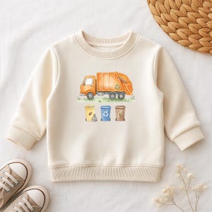 Könnte beinhalten: Cremefarbenes Sweatshirt mit einer Aquarellillustration eines orangefarbenen Müllwagens und drei Recyclingbehältern. Das Design umfasst gelbe, blaue und braune Behälter mit einem Recycling-Symbol auf dem LKW. Das Sweatshirt hat einen Rundhalsausschnitt und lange Ärmel.