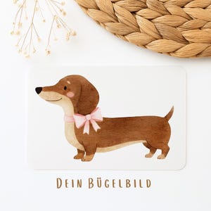 Könnte beinhalten: Aquarell-Illustration eines Dackelhundes mit braunem Fell und einer rosa Schleife. Der Hund steht auf weißem Hintergrund. Der Text "DEIN BÜGELBILD" ist braun am unteren Bildrand aufgedruckt.
