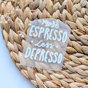 Könnte beinhalten: Ein klarer Aufkleber mit den Worten "more ESPRESSO less DEPRESSO" in weißer, kursiver Schrift. Der Aufkleber befindet sich auf einem gewebten, hellbraunen Platzset. Kleine weiße Blumen sind im Hintergrund.