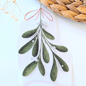 Könnte beinhalten: Eine Illustration von Mistelzweigen mit grünen Blättern und weißen Beeren, gebunden mit einer roten Schleife. Die Abbildung befindet sich auf weißem Hintergrund, mit einem Weidenkorb und Trockenblumen im Hintergrund.