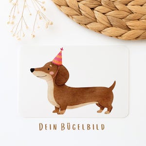 Könnte beinhalten: Aquarell-Illustration eines braunen Dackels mit Partyhut. Der Hund ist im Profil dargestellt, mit einem rosa Halsband. Der Text "DEIN BÜGELBILD" ist braun am unteren Bildrand aufgedruckt.