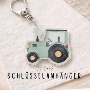 Könnte beinhalten: Ein klarer Acryl-Schlüsselanhänger mit einem grünen Traktormotiv, schwarzen Rädern und gelben Akzenten. Der Schlüsselanhänger hat einen silberfarbenen Metallverschluss. Der Text "SCHLÜSSELANHÄNGER" ist unten im Bild in Schwarz gedruckt.