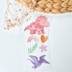 Könnte beinhalten: Ein Stickerbogen mit einem rosa Triceratops mit Herzmustern, einem Regenbogen, einem orangefarbenen Herz, einem grünen Blatt, einer lila Blume und einem lila Pterodactylus. Die Aufkleber befinden sich auf weißem Hintergrund.