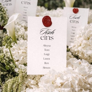 May include: A white rectangular card with a textured surface featuring the black printed text Tisch eins and a list of names including Aleyna, Tom, Luigi, Laura, Ben, and Simon. A dark red wax seal with a floral design is centered at the top.