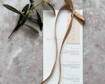Puede incluir: Una tarjeta de menú blanca, alta y rectangular, con texto en caligrafía elegante, atada con una cinta de satén dorada larga y fina. Una pequeña etiqueta rectangular de color beige con los nombres Fiona & Henry escritos a mano cuelga del lazo.
