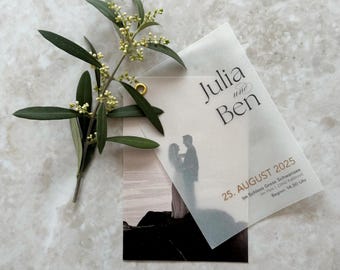 Puede incluir: Una invitación de boda con los nombres "Julia und Ben" impresos en una fuente elegante. La invitación presenta una silueta de una pareja y la fecha "25. AUGUST 2025". Una ramita de ramas de olivo con pequeñas flores blancas añade un toque natural.