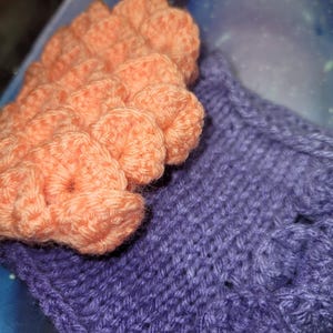 May include: A close-up of a crocheted hat with a purple and peach color scheme. The hat is made with a textured stitch and has a ruffled edge.