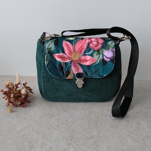 Può includere: Borsa a tracolla in velluto a coste verde acqua con patta con stampa floreale rosa, rossa e viola. La borsa ha una tracolla nera regolabile e una chiusura color argento. Un piccolo arrangiamento di fiori secchi è a sinistra.