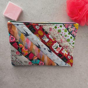 Peut inclure: Une pochette en tissu matelassé avec un motif à rayures diagonales colorées. Le motif comprend des motifs floraux, de chats porte-bonheur et d'oiseaux. La pochette a une fermeture éclair avec une tirette arc-en-ciel. Un pain de savon rose et une fleur de douche rose sont également dans l'image.