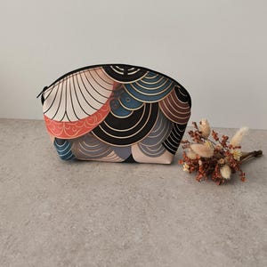 Peut inclure: Une trousse de maquillage colorée avec une fermeture éclair. Le sac présente un motif de formes superposées dans des tons de rouge, noir, bleu et marron. Le sac est posé sur une surface grise.