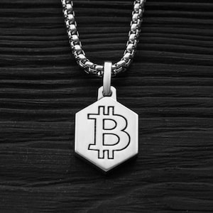 May include: Silver-toned necklace with a hexagonal pendant featuring the Bitcoin symbol. The pendant hangs from a chain with a textured, rounded link design. The background is a dark, textured wooden surface.