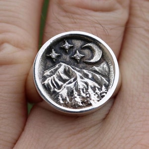 Peut inclure: Une bague en argent avec un motif détaillé d'une chaîne de montagnes, d'un croissant de lune et de trois étoiles. La bague est portée au doigt d'une personne.