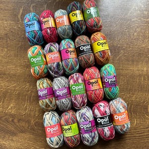 May include: A collection of Opal brand yarn skeins in various colors, including teal, red, and green. Each skein has a colorful, variegated pattern and is wrapped with a label. The yarn is arranged on a wooden surface.