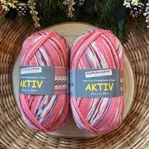 Könnte beinhalten: Zwei Knäuel Aktiv-Garn in Rosa, Weiß und Blau, mit einem Etikett "SUPERGARNE". Jedes Knäuel wiegt 100 g und hat eine Lauflänge von ca. 420 Metern. Das Garn liegt in einem Weidenkorb.