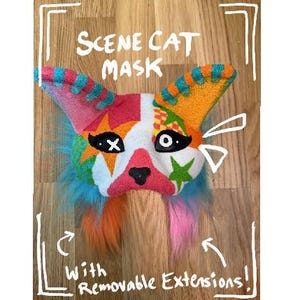 May include: A colorful Scene Cat mask with removable extensions. The mask features a white base with a star-shaped design around the eyes and a pink nose. The ears are striped with blue, orange, and pink. The mask has blue, orange, and pink fur extensions.