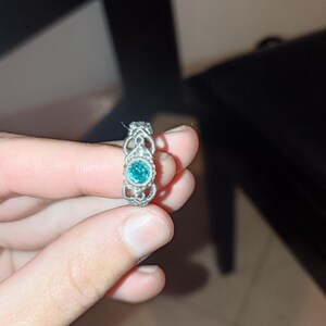 May include: A silver ring with an ornate design. The ring features a round, teal-colored gemstone at its center, surrounded by a detailed filigree pattern. The ring is held up by a person's fingers.