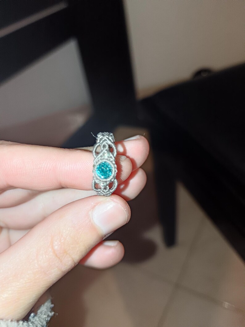May include: A silver ring with an ornate design. The ring features a round, teal-colored gemstone at its center, surrounded by a detailed filigree pattern. The ring is held up by a person's fingers.