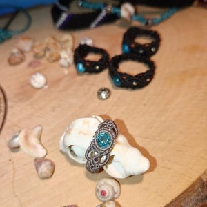 May include: A handmade macrame ring with a light blue gemstone center, displayed on a seashell. Several other macrame rings and bracelets in black and blue are visible in the background, along with small seashells.