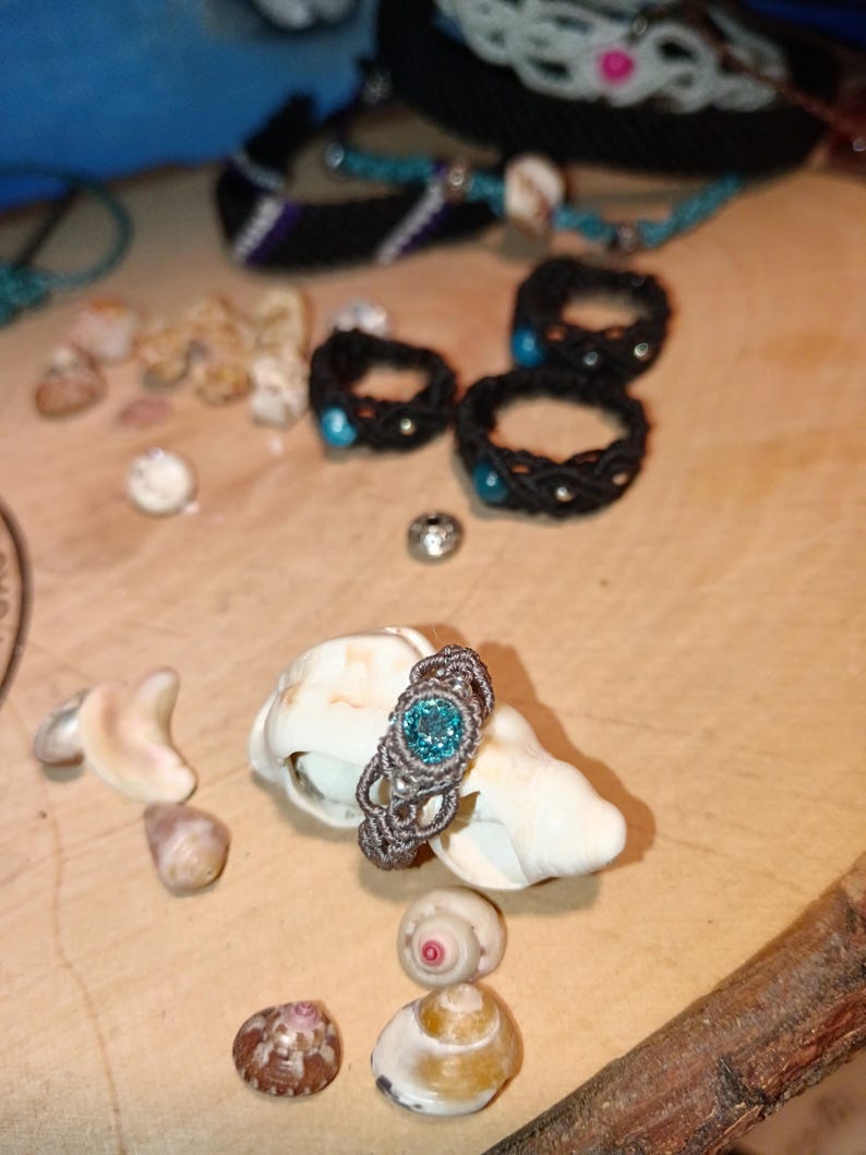 May include: A handmade macrame ring with a light blue gemstone center, displayed on a seashell. Several other macrame rings and bracelets in black and blue are visible in the background, along with small seashells.